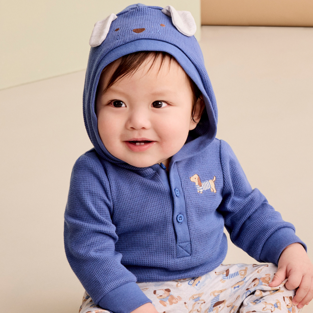 A baby wearing a blue waffle-knit hooded long-sleeve top with white bunny ears on the hood and a small dog graphic on the chest, paired with patterned pants, sitting against a soft beige background.