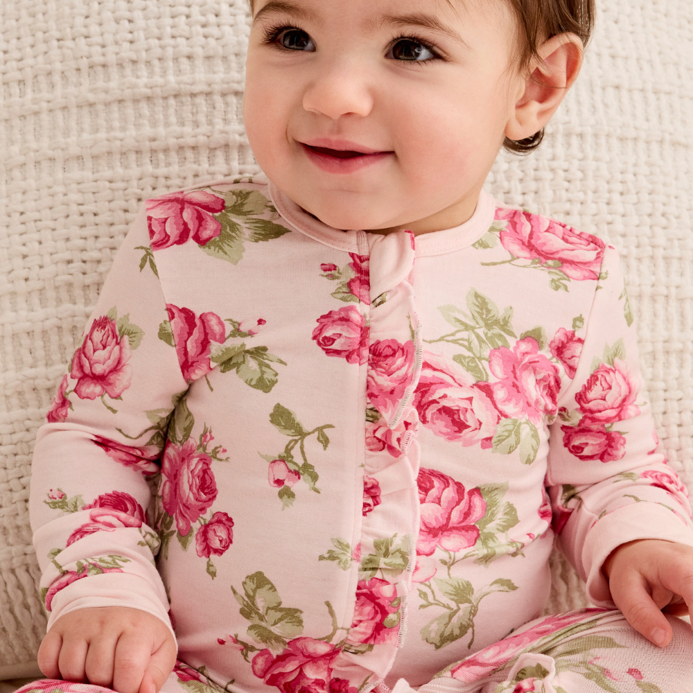 A baby wearing a light pink long-sleeve footless sleeper with a pink rose and green leaf floral print, sitting on a textured cream knit blanket.