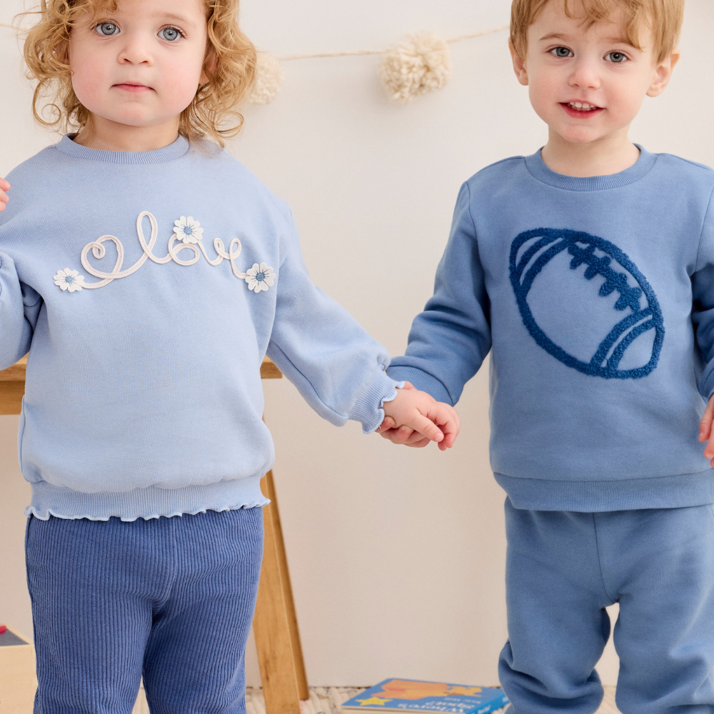 Two young children standing indoors and holding hands, wearing matching light blue sweatshirt and pants sets. The child on the left has white embroidered script and flowers on the chest, and the child on the right has a dark blue football graphic on the chest.
